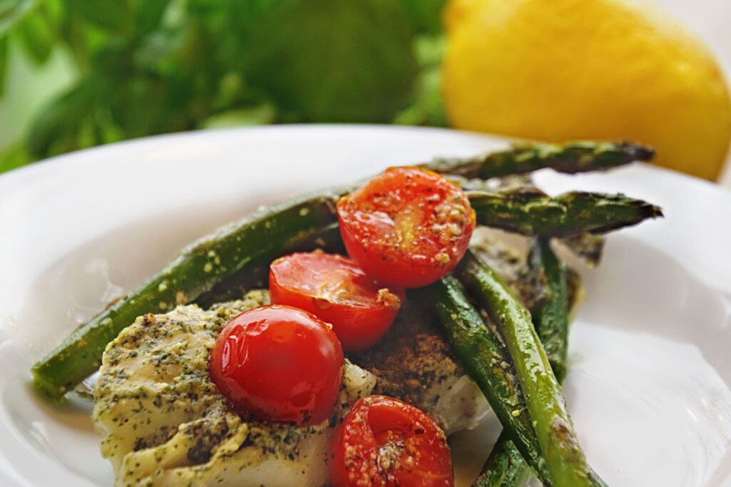 baked cod with pesto cherry tomatoes and asparagus on a white plate and lemon and basil in background.