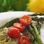baked cod with pesto cherry tomatoes and asparagus on a white plate and lemon and basil in background.