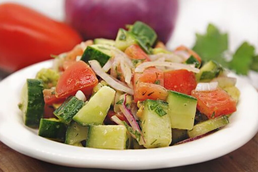greek salad with cucumber, tomato and red onion in white bowl with red onion and tomato in background.