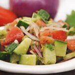 greek salad with cucumber, tomato and red onion in white bowl with red onion and tomato in background.