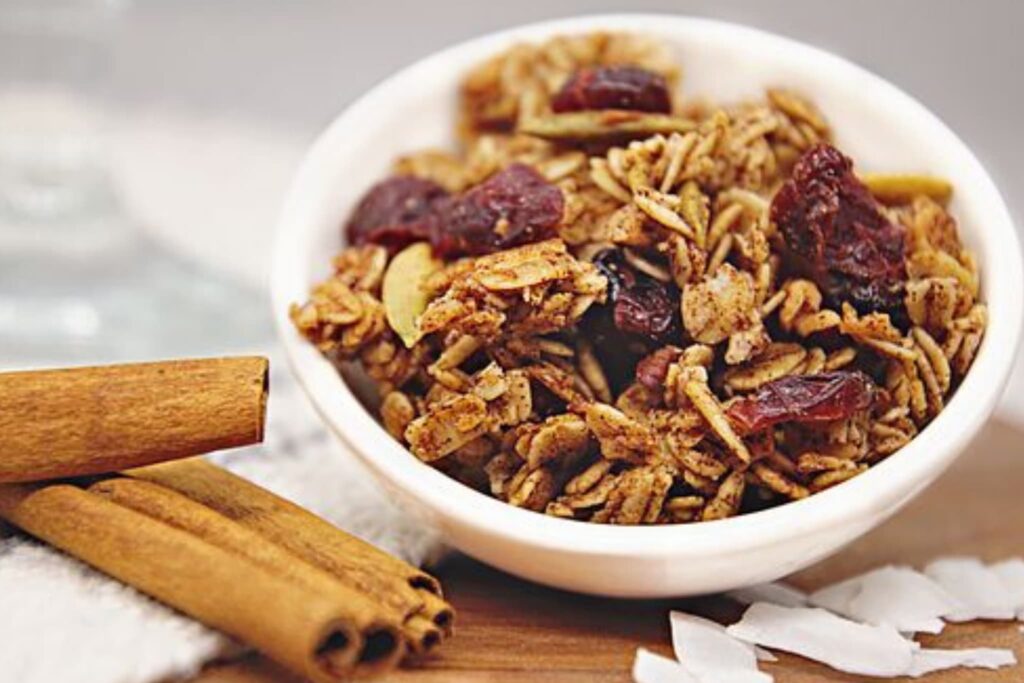 Granola with oats, dried cherries and seeds in white bowl on board and cinnamon sticks and shaved coconut.