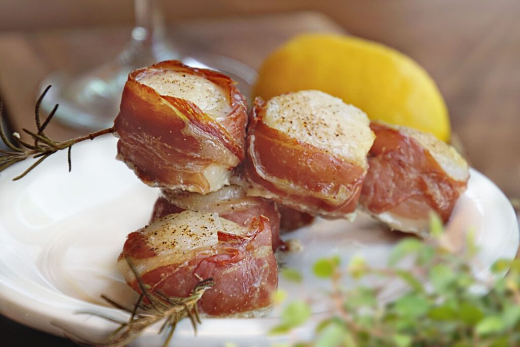 prosciutto wrapped scallops with rosemary sprigs on white plate with herbs and lemon in background.
