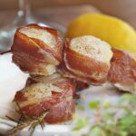 prosciutto wrapped scallops with rosemary sprigs on white plate with herbs and lemon in background.