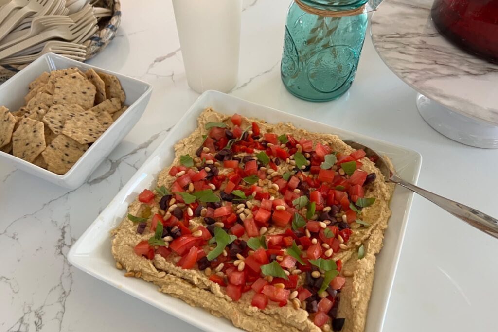 Hummus on white platter topped with tomatoes olives and basil.